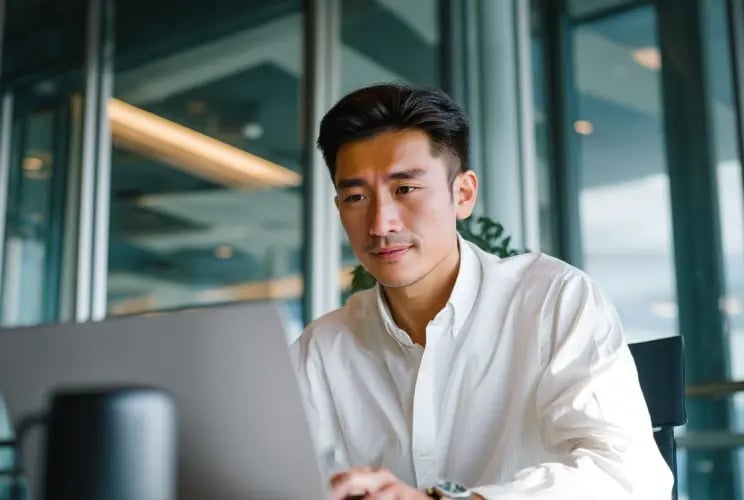 Young Asian professional in white shirt working on laptop in modern glass office environment