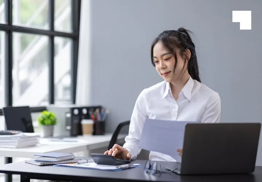 Professional Asian businesswoman reviewing financial documents at modern office desk with laptop and calculator